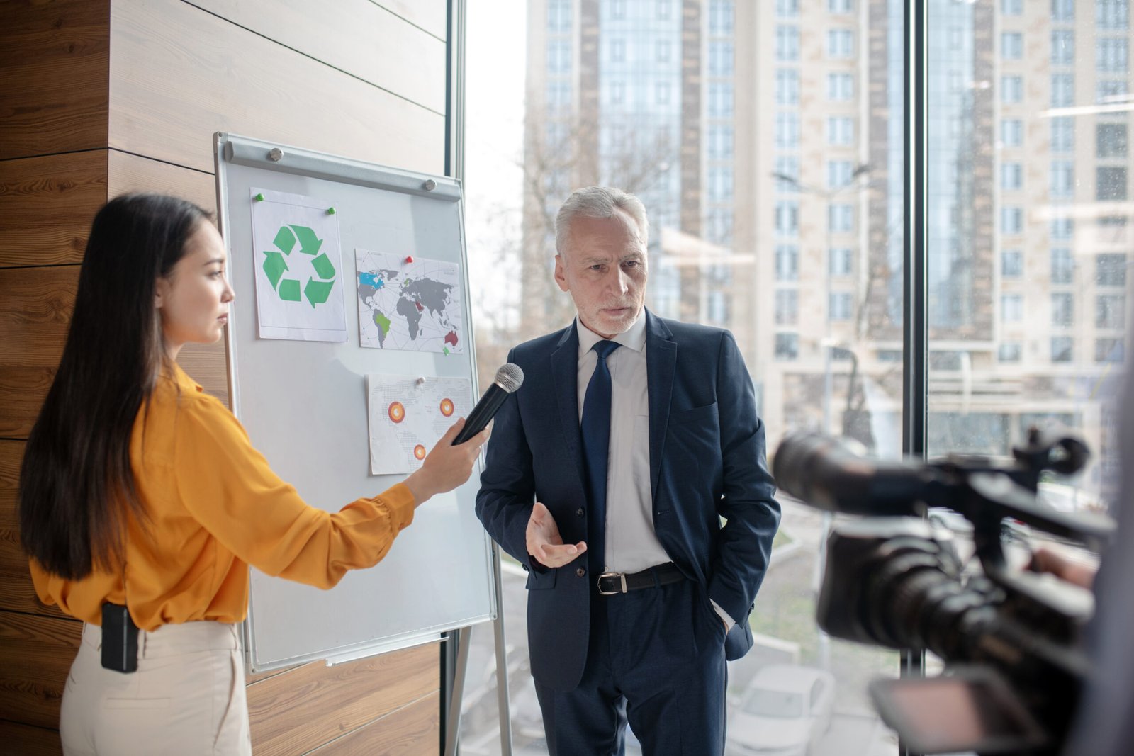 During interview. Dark-haired female reporter holding a microphone while standing near the white board